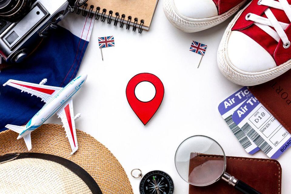 A flat-lay of travel planning items around a red map pin: a passport, air tickets, a camera, notebook, compass, sun hat and trainers.