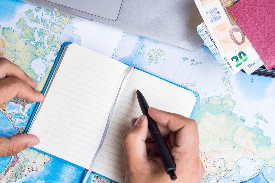 A traveller writing in a notebook laid over a world map, with euro banknotes, a passport and a laptop beside them.