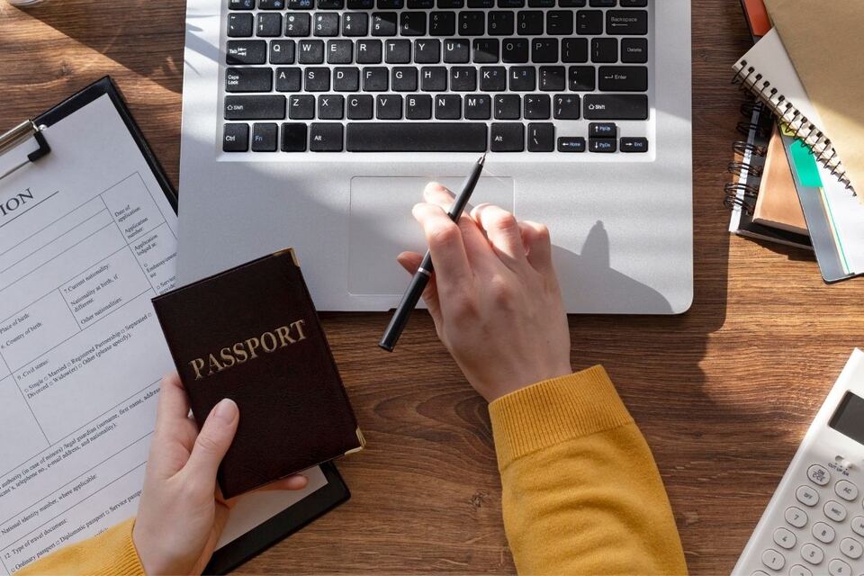 An overhead view of someone holding a passport beside a laptop, a blank visa application form, a notebook and a calculator on a wooden desk.
