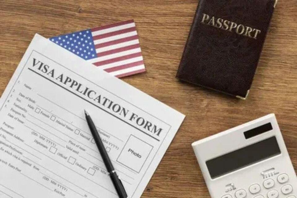A blank visa application form, passport, small flag, pen and calculator arranged on a wooden desk.
